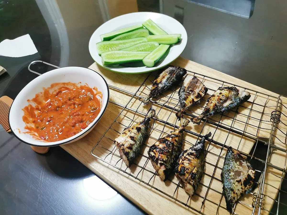 Pescado a la parrilla sobre una rejilla con cuenco de sambal rojo y rodajas de pepino