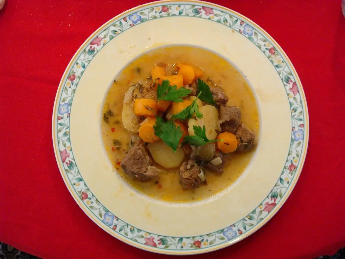 Plato con guiso de cordero, patata y zanahoria en caldo claro con perejil sobre un mantel rojo
