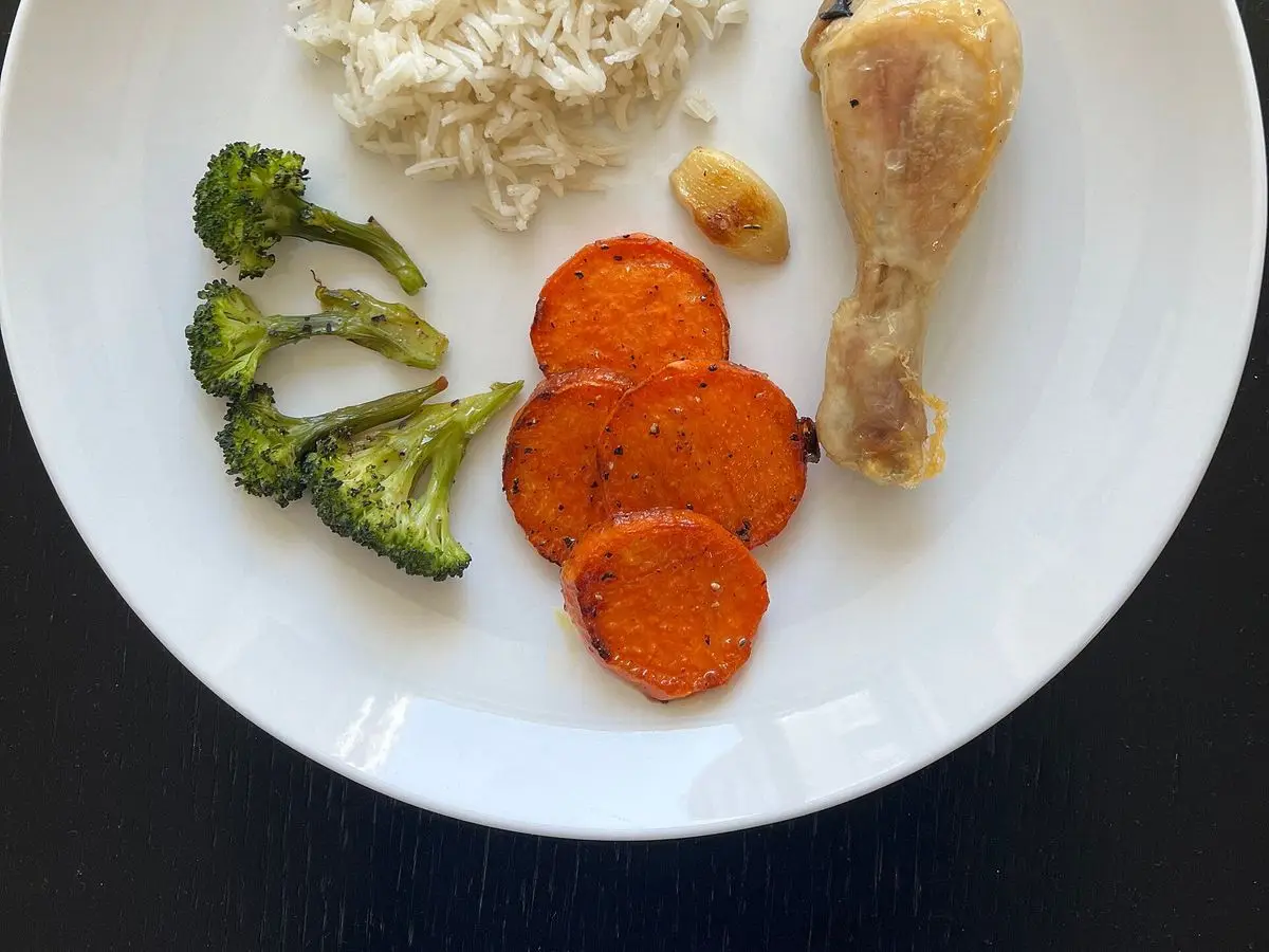 Plato blanco con muslito de pollo asado, arroz blanco, brócoli y rodajas de zanahoria