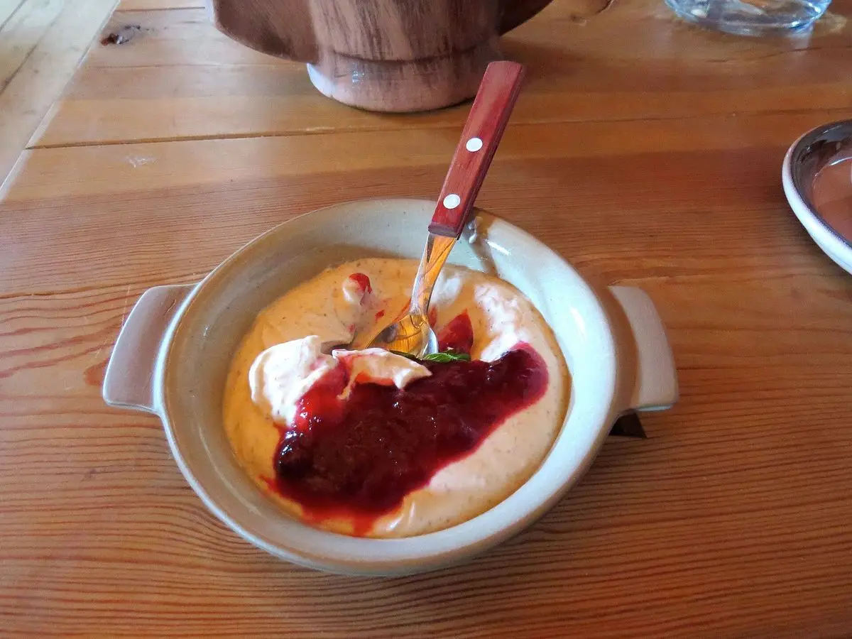 Cuenco con postre cremoso de kama, nata y mermelada roja con una cuchara sobre una mesa de madera