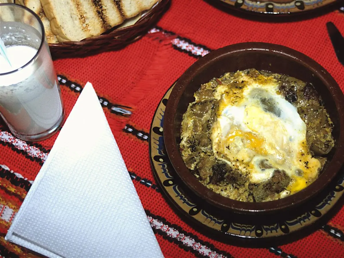 Cazuela de barro con guiso búlgaro de carne cubierto de huevo y yogur, sobre un mantel rojo