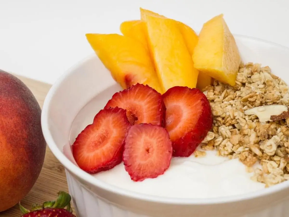 Cuenco blanco de yogur con gajos de melocotón, fresas y granola de avena con almendra, junto a un melocotón entero