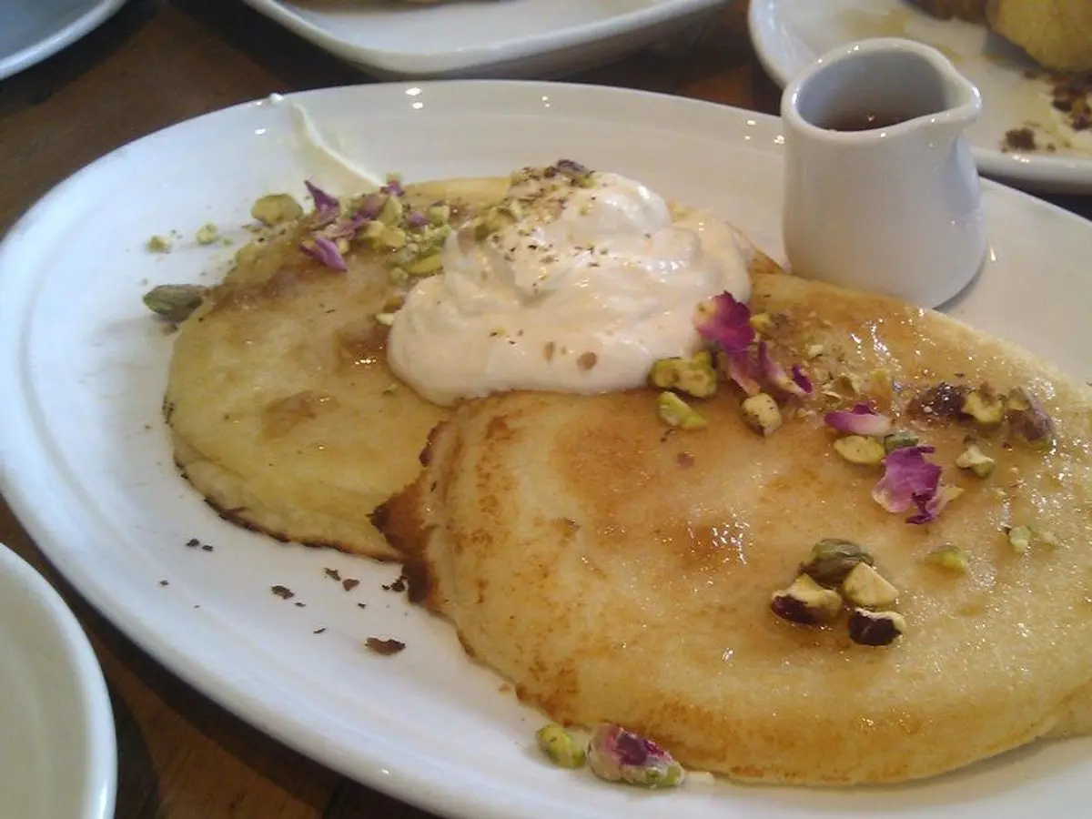 Dos tortitas doradas bañadas en almíbar con pistachos, pétalos de rosa y crema, junto a una jarrita de sirope