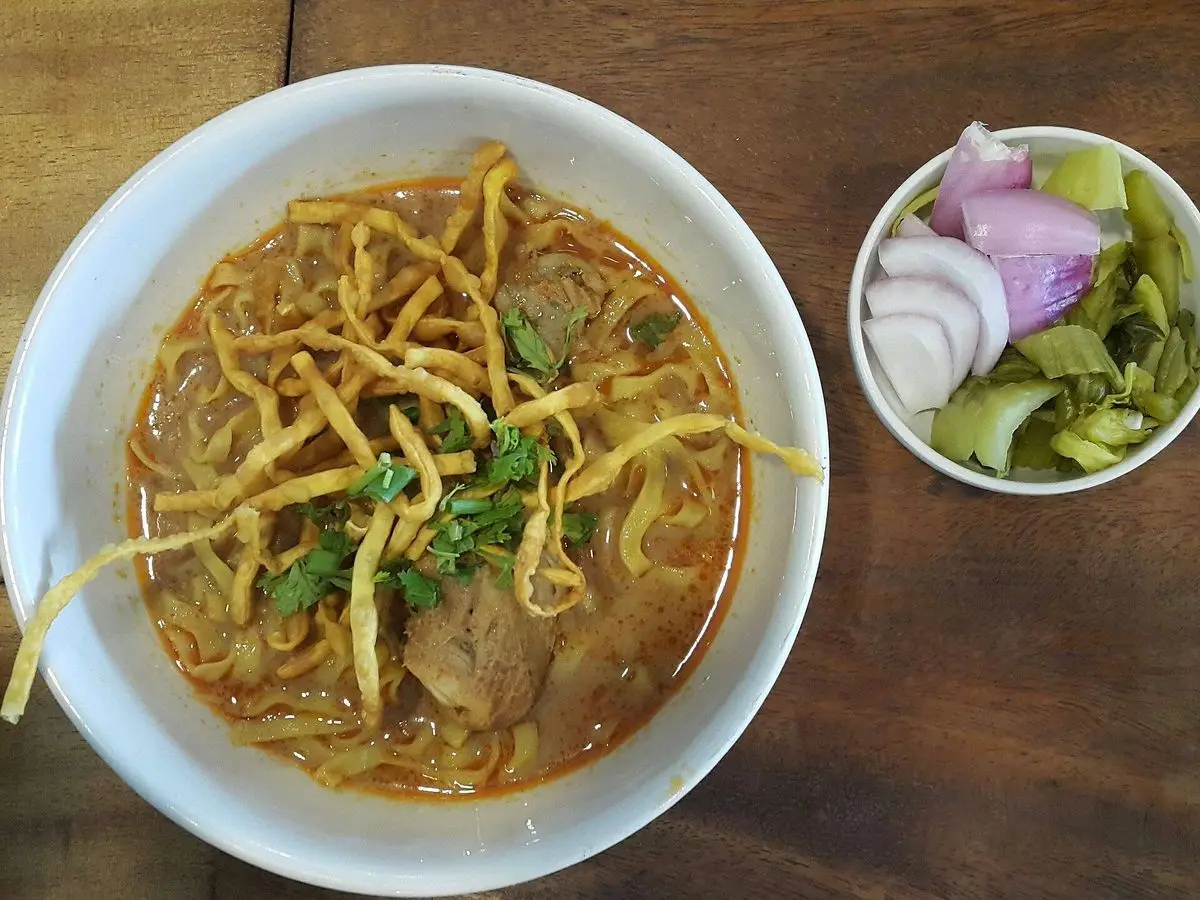 Cuenco blanco de sopa de curry con fideos fritos crujientes por encima, con cebolla morada y verdura a un lado