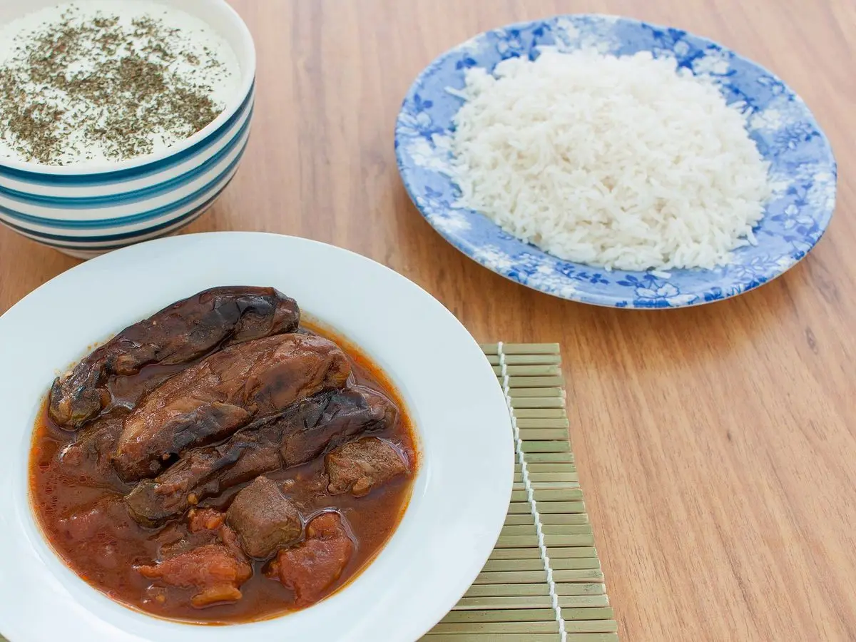 Plato de guiso persa de berenjena en salsa de tomate junto a un plato de arroz blanco sobre una mesa de madera
