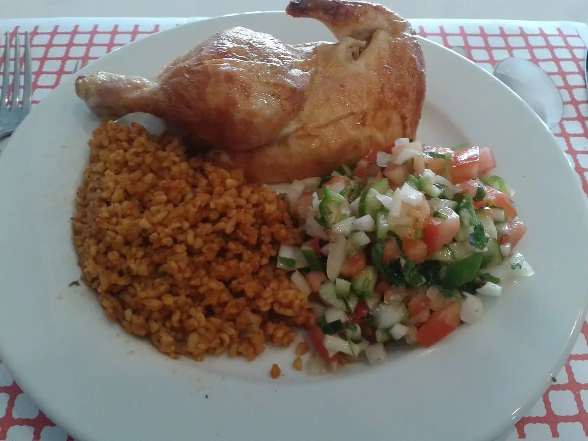 Plato con pollo asado, bulgur anaranjado y ensalada fresca de tomate y pepino sobre mantel de cuadros