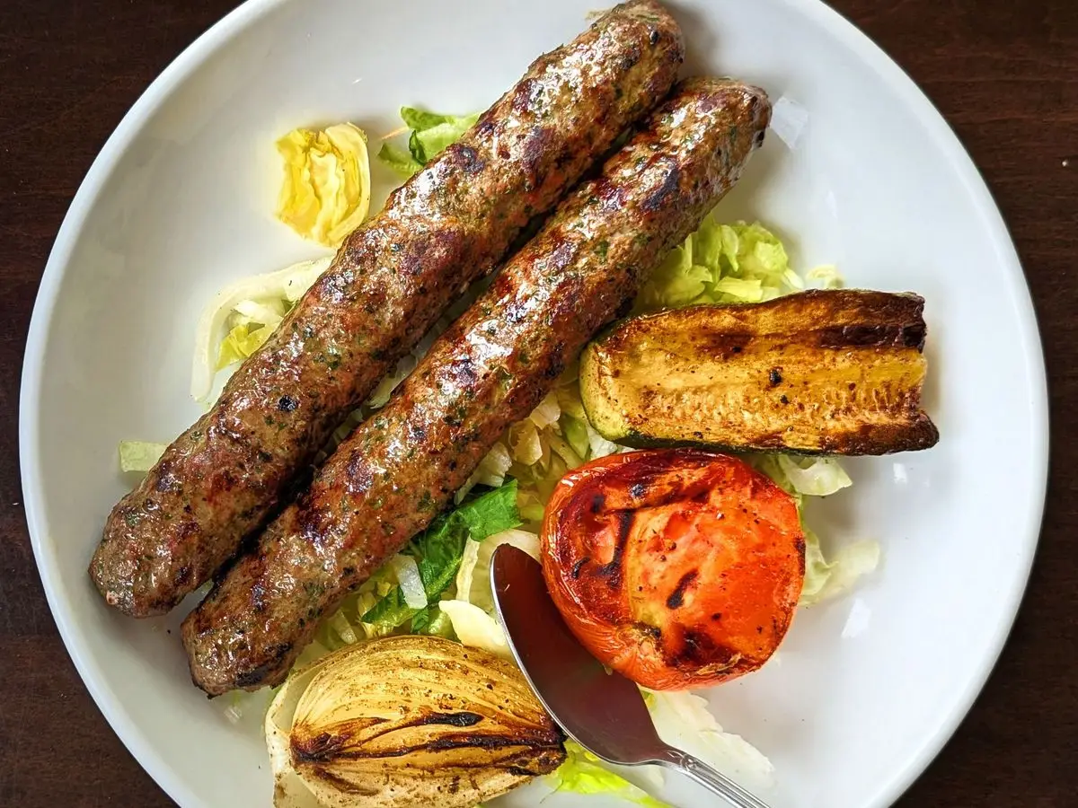 Dos brochetas de carne picada con tomate y cebolla asados sobre ensalada en un plato blanco