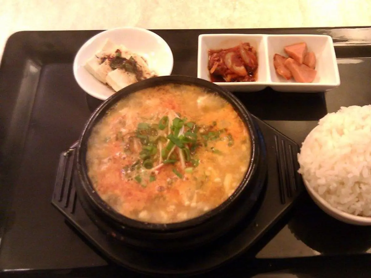 Cazuela negra de guiso coreano de tofu con verduras, junto a un cuenco de arroz y encurtidos