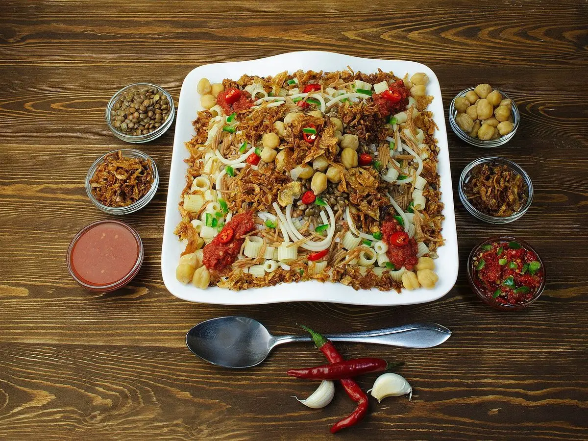 Fuente blanca de koshari con arroz, lentejas, pasta, garbanzos y cebolla frita rodeada de cuencos de salsas