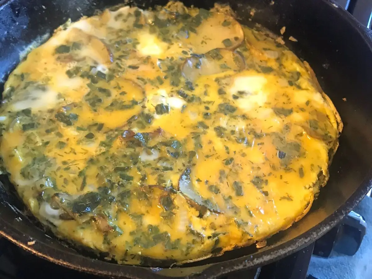 Tortilla de hierbas verdes dorada cuajándose en una sartén negra, kuku sabzi