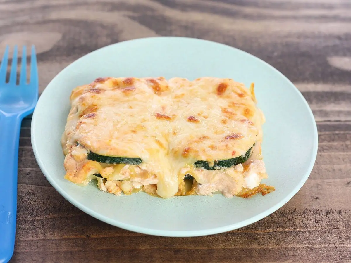 Ración de lasaña de calabacín gratinada con queso en un plato verde sobre una mesa de madera