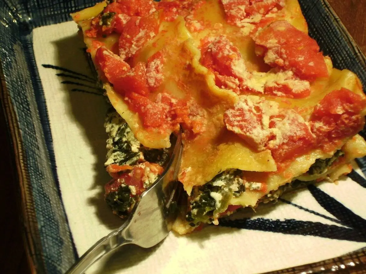Porción de lasaña vegetal con espinacas y salsa de tomate en una fuente, con un tenedor