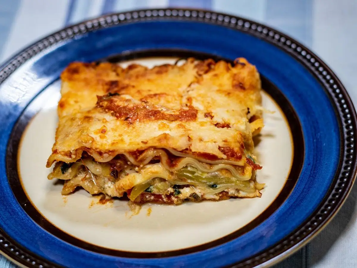 Porción de lasaña de verduras gratinada sobre un plato azul, con las capas visibles