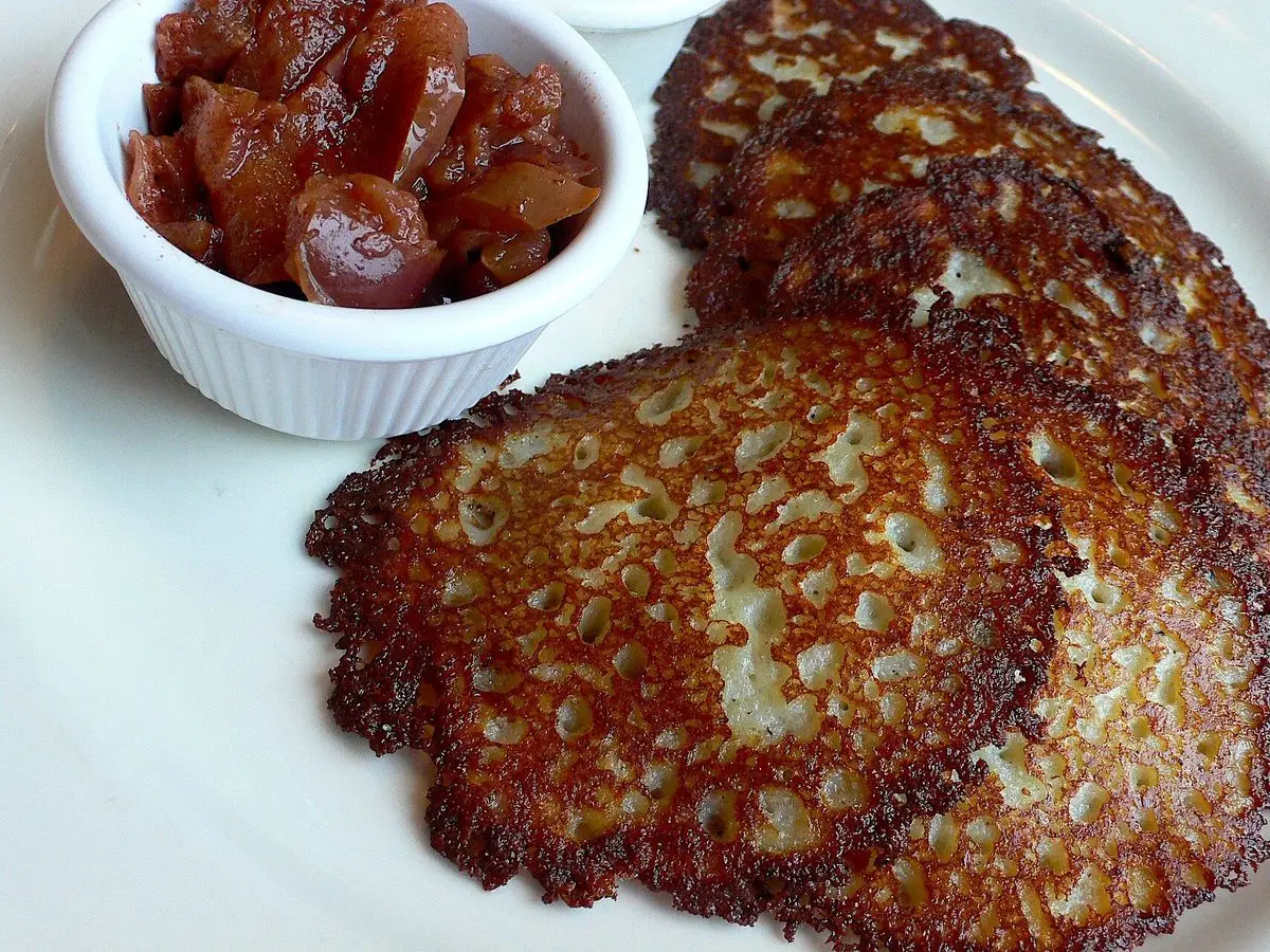 Latkes de patata dorados apilados en un plato blanco junto a un cuenco de compota de fruta