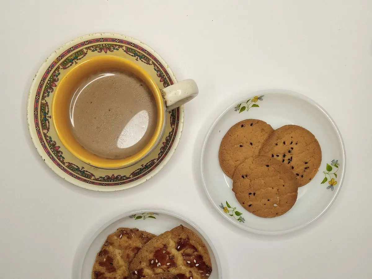 Taza de café con leche junto a un plato de galletas sobre una mesa blanca