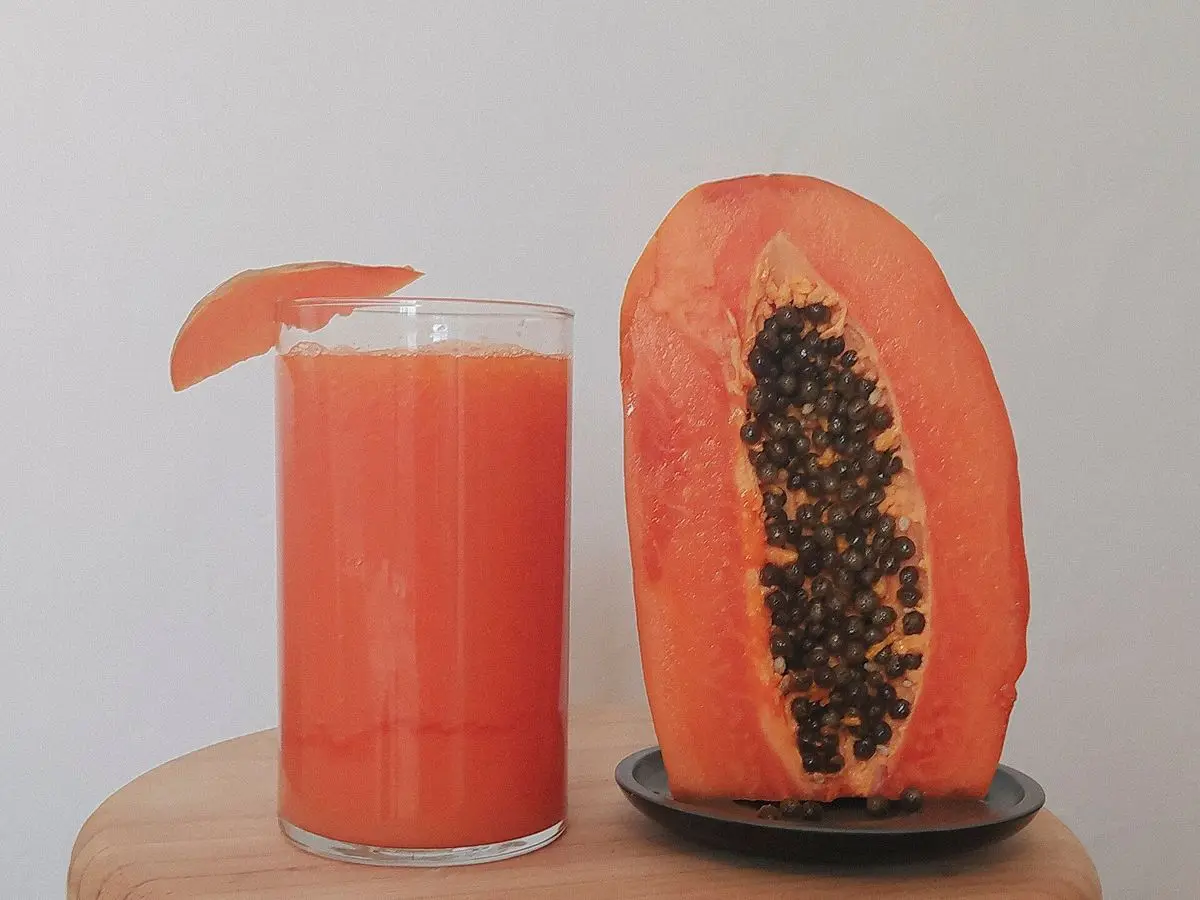 Vaso de licuado de papaya anaranjado junto a una papaya abierta con semillas negras sobre una tabla