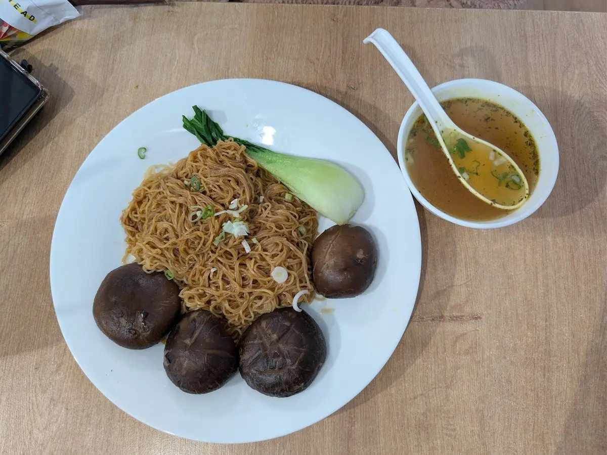 Plato blanco de fideos salteados con setas shiitake y pak choi junto a un cuenco de caldo