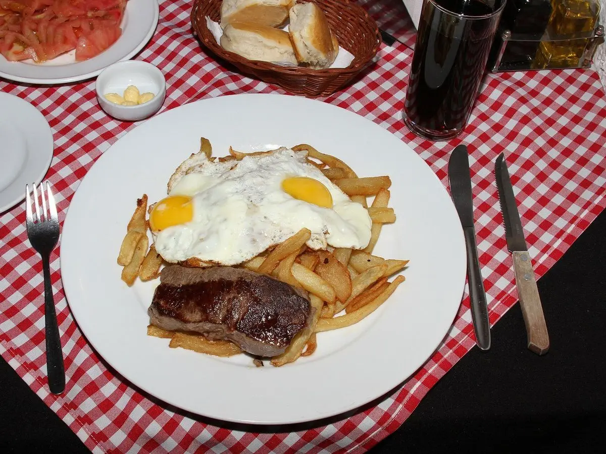 Plato de filete de ternera con patatas fritas, cebolla y huevo frito sobre mantel de cuadros