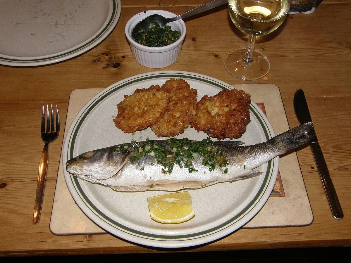 Lubina entera cocinada con hierbas y limón en un plato ovalado junto a una guarnición dorada