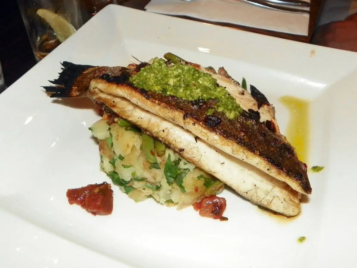 Lomo de lubina a la plancha con salsa verde sobre patata y verduras en un plato cuadrado blanco