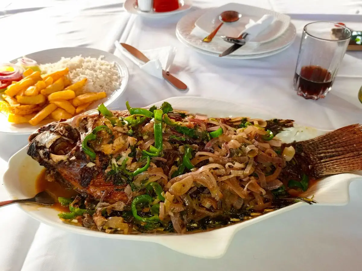 Pescado entero cubierto de cebolla y pimiento verde, con patatas fritas y arroz al fondo