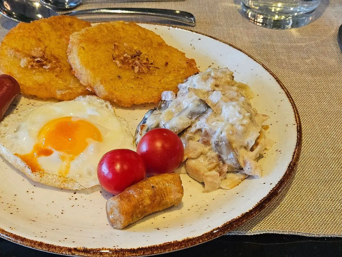 Plato con tortitas de patata doradas, salsa cremosa con carne, huevo frito, salchichas y tomates cherry