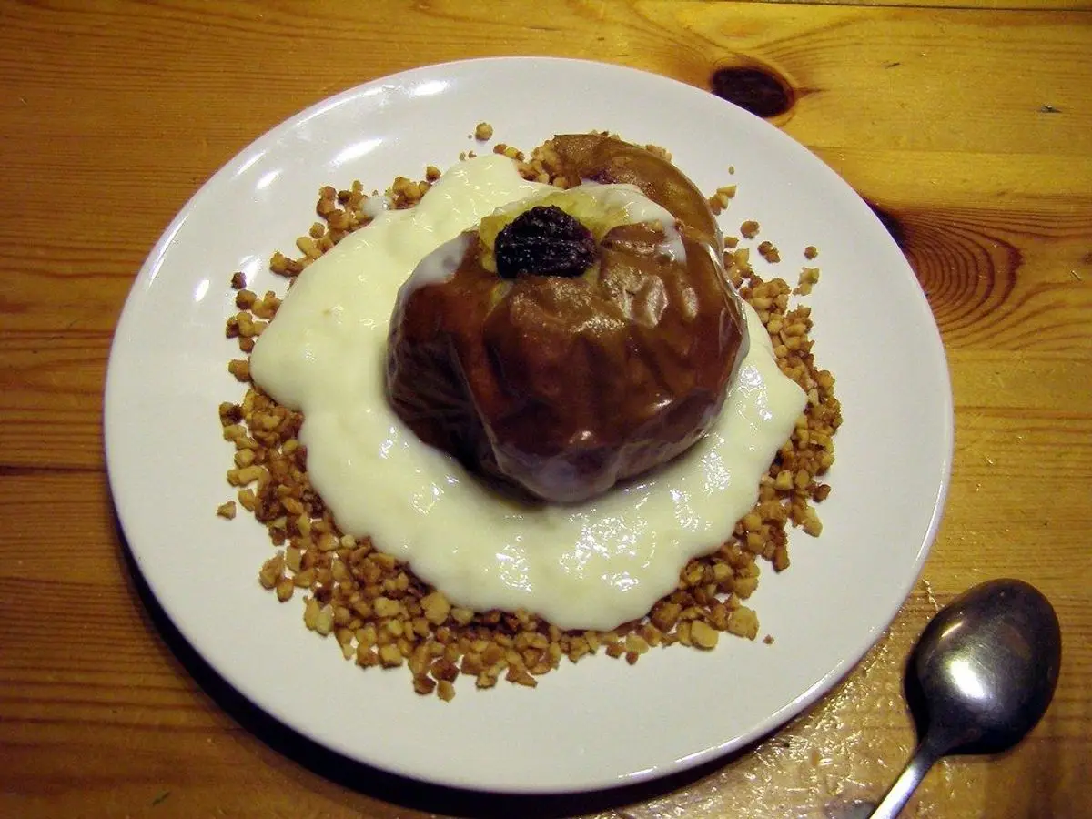 Manzana asada sobre una capa de crema blanca rodeada de frutos secos picados en un plato blanco