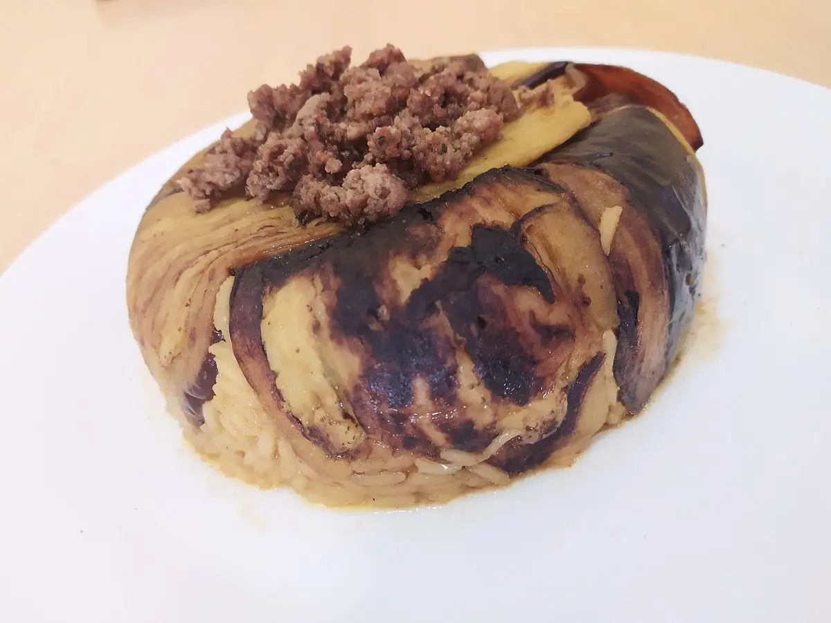 Maqluba volteada envuelta en berenjena asada y coronada con carne picada dorada sobre un plato blanco