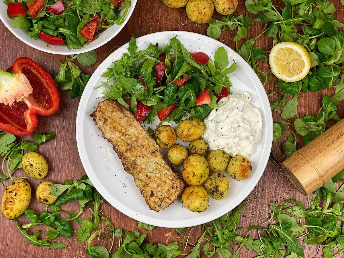 Filete de pescado al horno con patatas pequeñas, ensalada verde con tomate y limón sobre una mesa de madera
