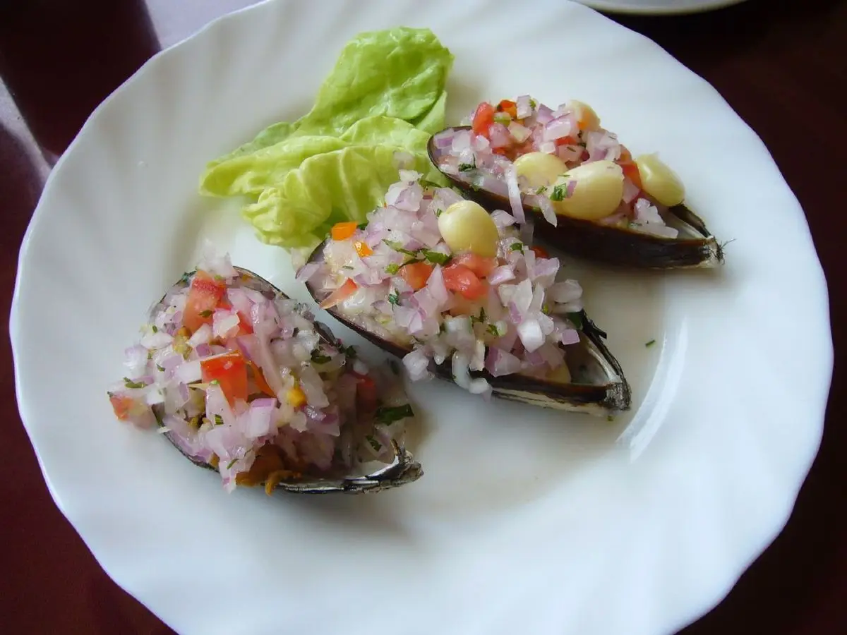 Mejillones en su concha cubiertos con una vinagreta de cebolla, tomate y pimiento en un plato blanco con lechuga