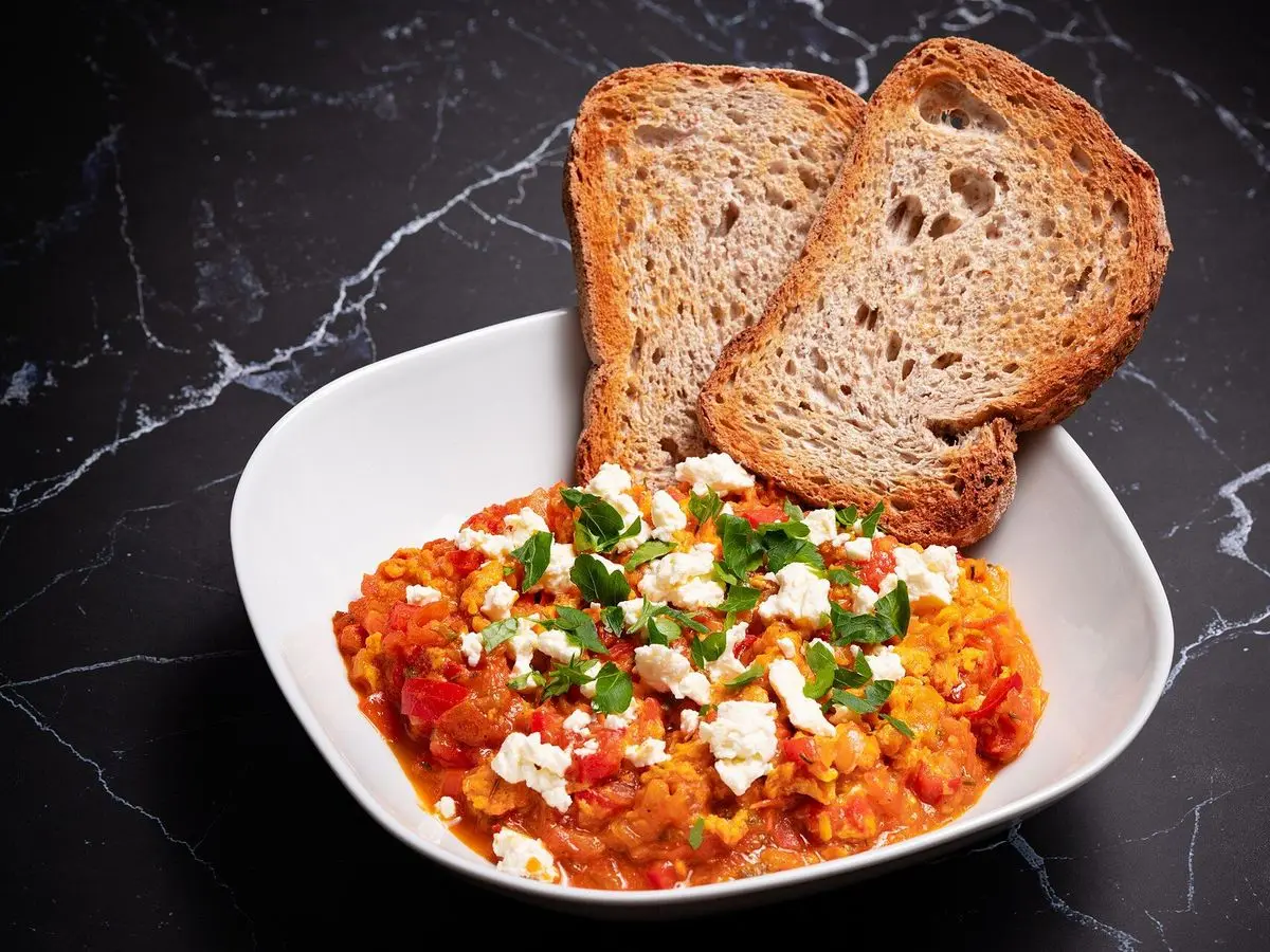 Cuenco blanco de menemen, huevo revuelto con tomate y pimiento, con queso blanco, hierbas y dos rebanadas de pan