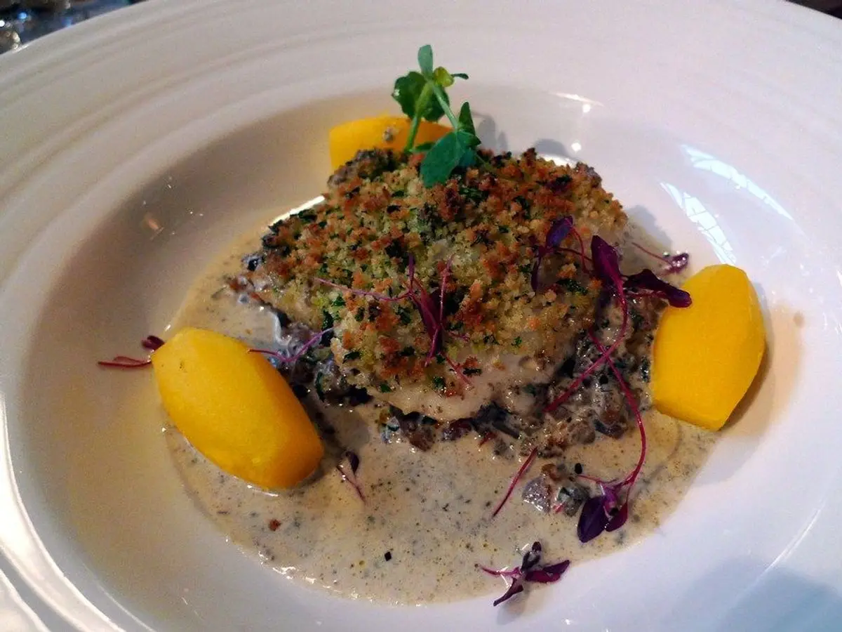 Pescado blanco al horno con costra de hierbas, patatas amarillas y salsa cremosa en un plato hondo blanco