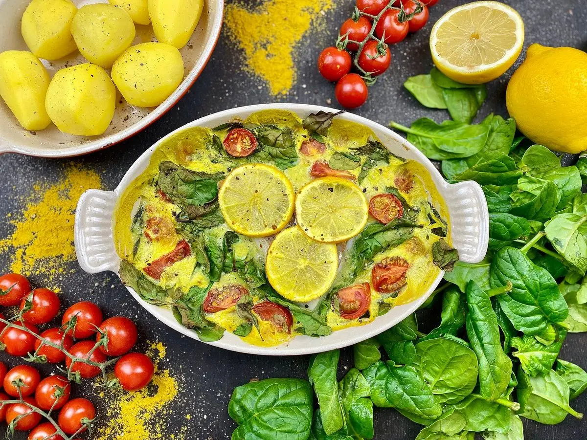 Pescado al horno sobre espinacas con rodajas de limón y tomatitos en una fuente blanca, rodeada de patatas y tomates