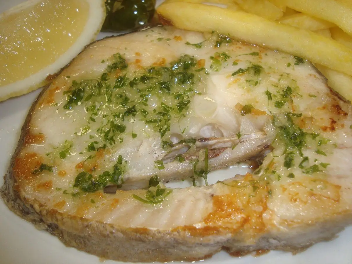 Rodaja de merluza a la plancha con perejil picado, medio limón y patatas fritas al fondo