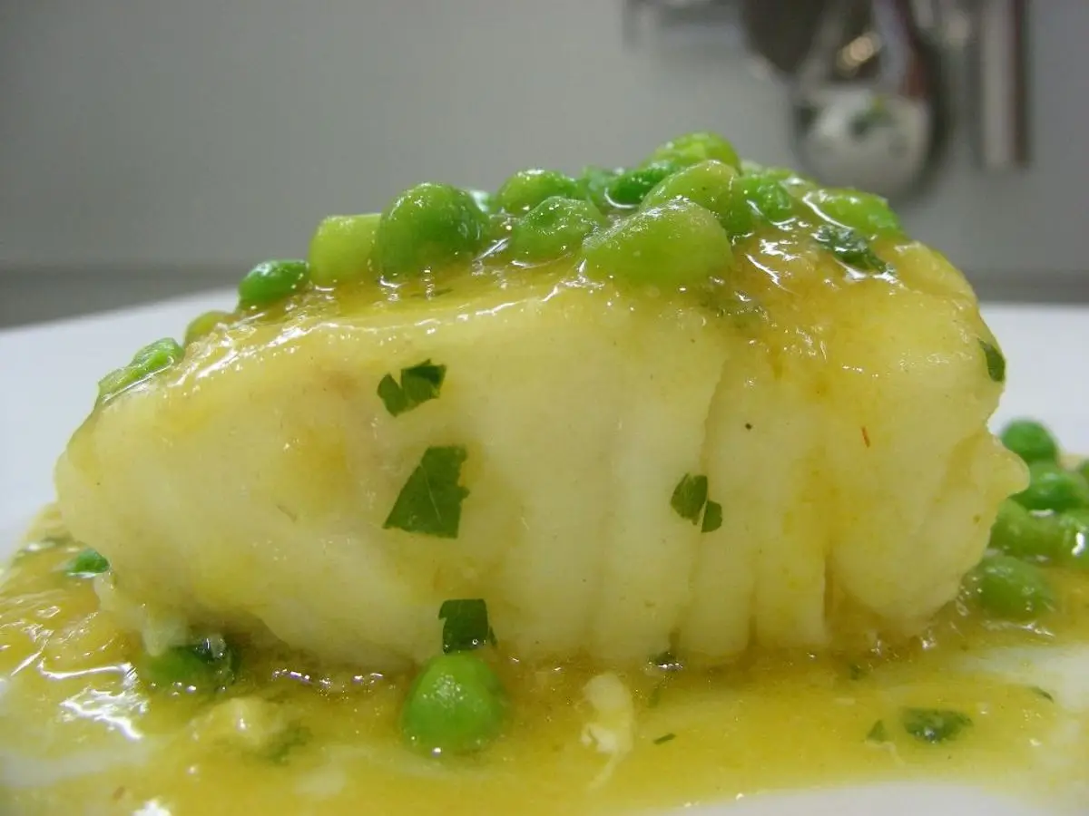Lomo de merluza cubierto de salsa ligada con guisantes y perejil picado en un plato blanco