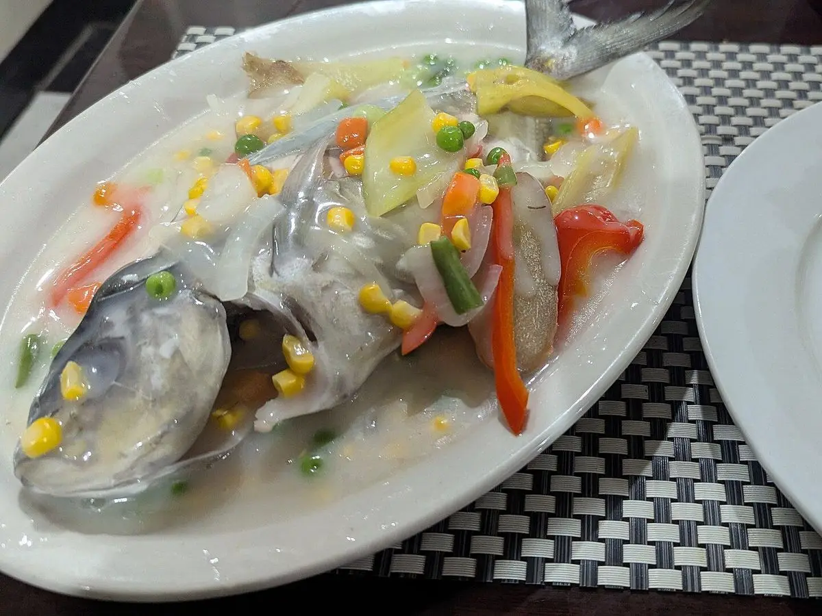 Pescado entero al vapor con maíz, pimiento rojo y verduras en una fuente blanca