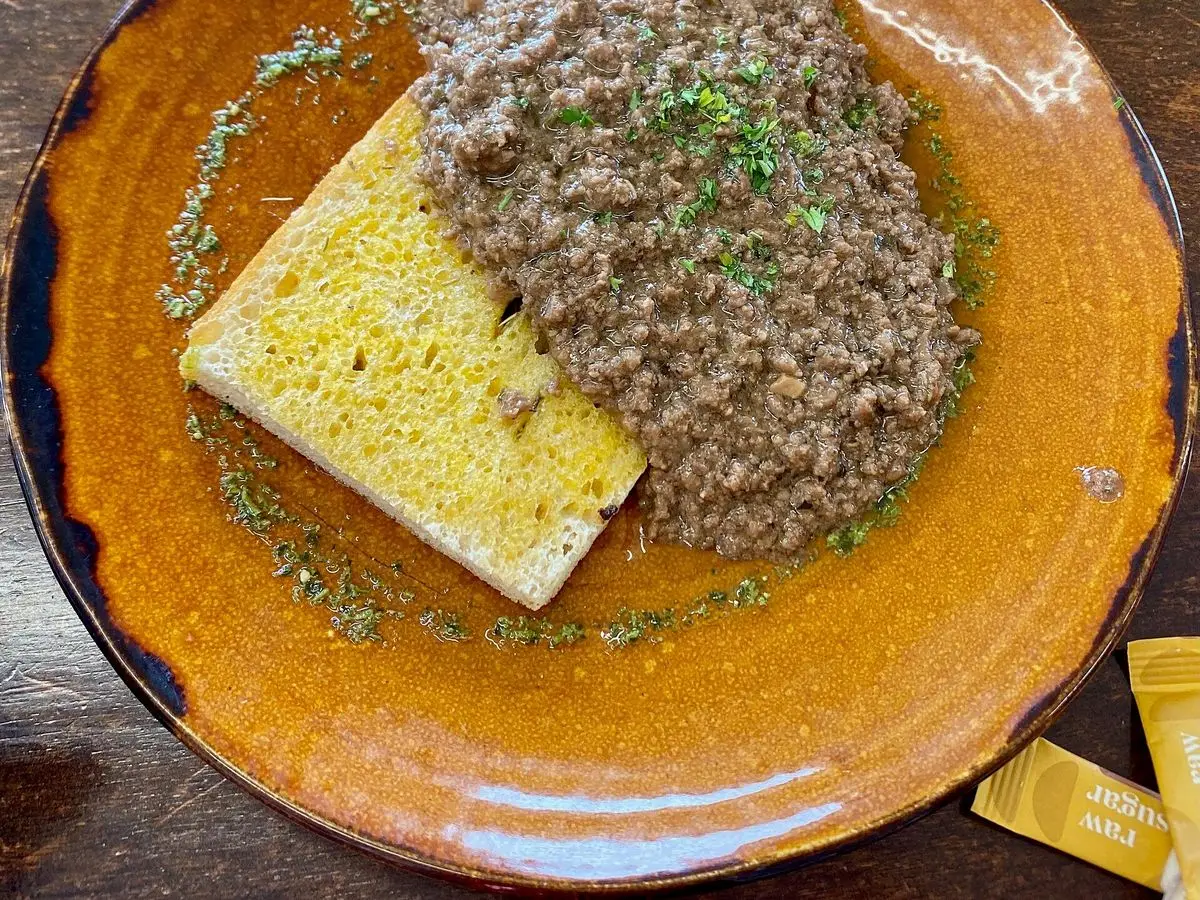 Plato con carne picada guisada sobre una tostada de pan, con un chorrito de salsa y hierbas