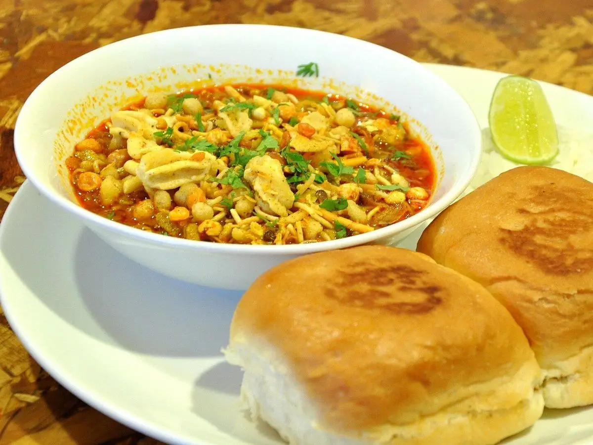 Cuenco de misal, curry especiado de legumbres con cebolla y cilantro, junto a dos panecillos pav y una rodaja de lima