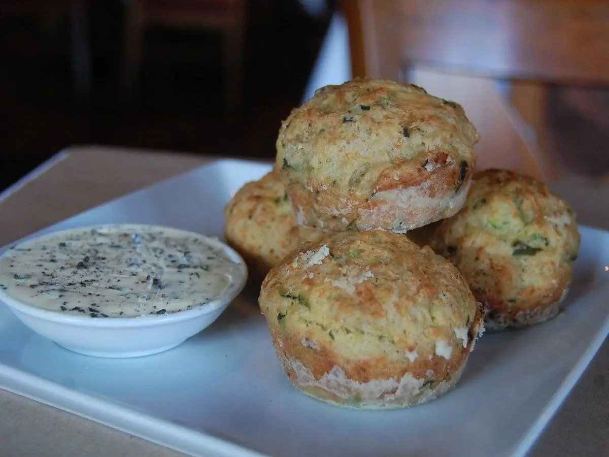 Muffins salados de calabacín dorados sobre un plato blanco junto a un cuenco con salsa