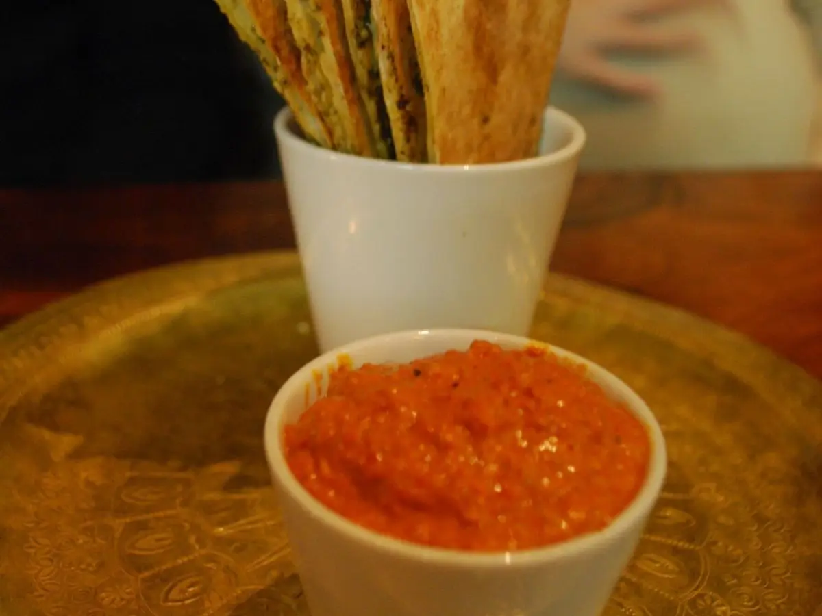 Vasito de crema roja de pimiento asado junto a un vaso con palitos de pan crujientes