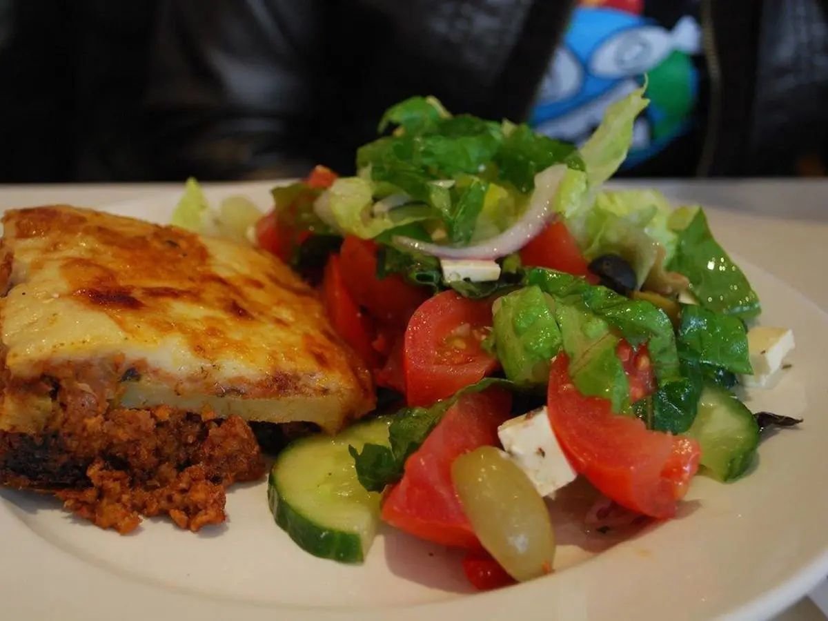 Porción de musaca gratinada junto a una ensalada de tomate, pepino y lechuga en un plato blanco