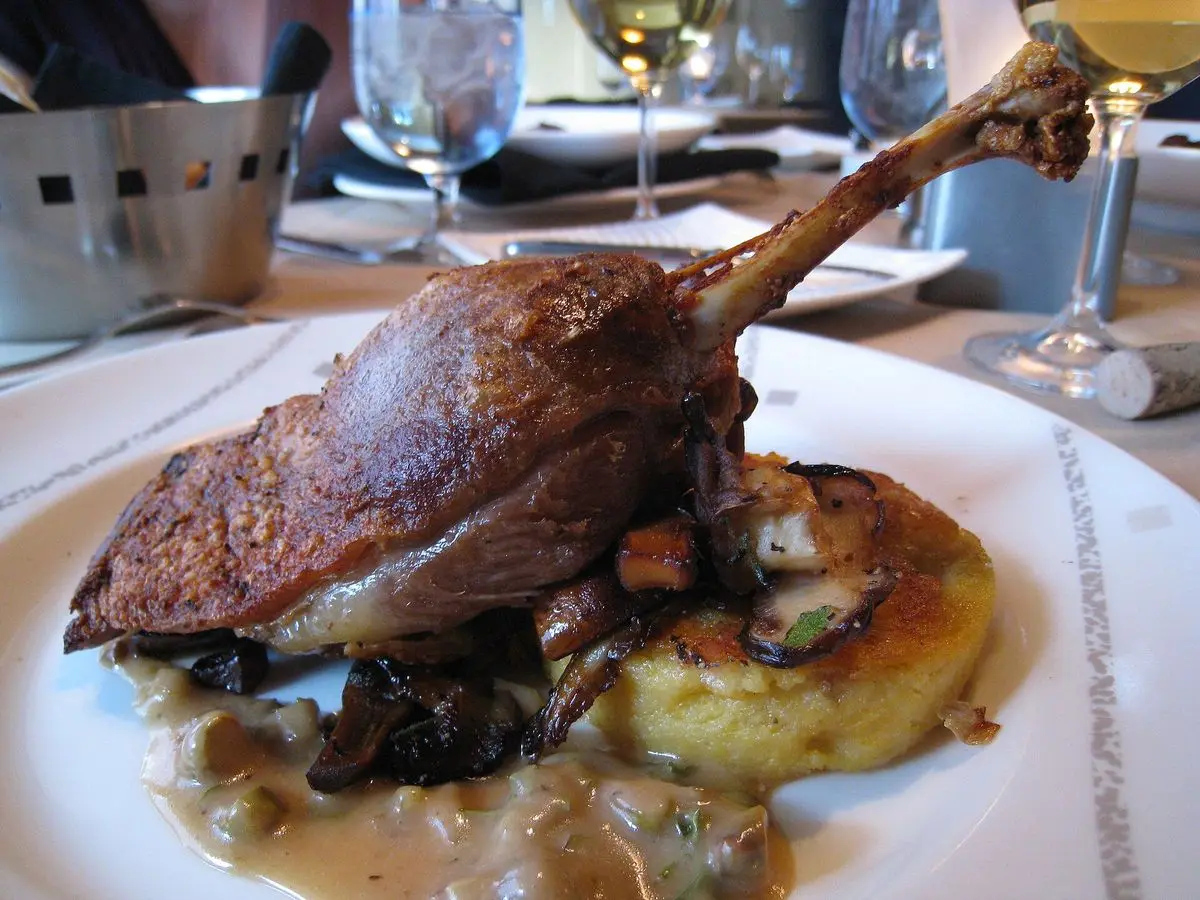 Muslo de pato confitado sobre una base de patata con salsa y setas en un plato blanco