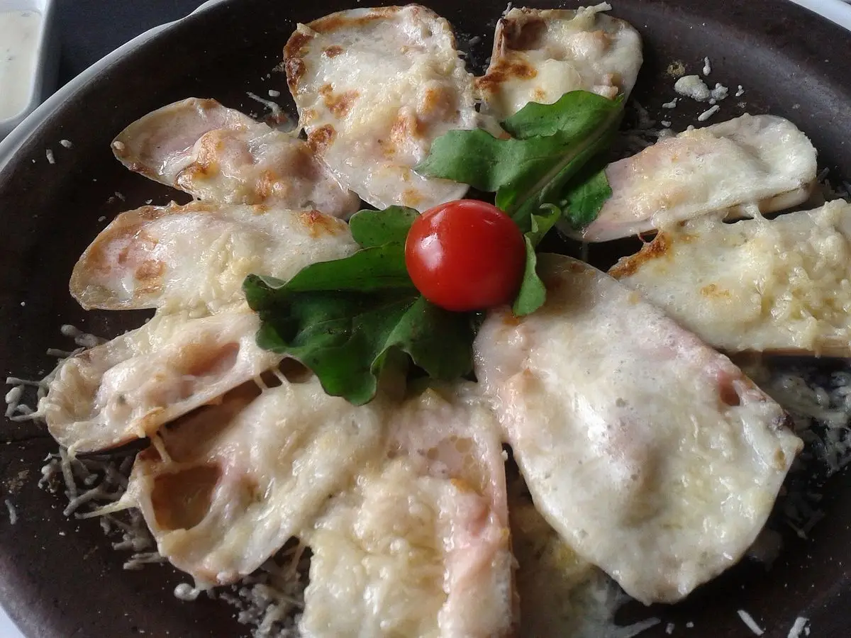 Machas gratinadas con queso parmesano en plato oscuro, con hojas de albahaca y un tomate cherry en el centro