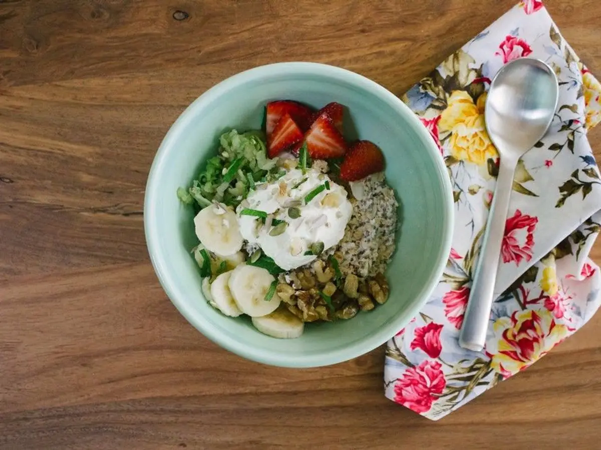 Cuenco verde de bircher con fresas, plátano, yogur y semillas visto desde arriba con una cuchara