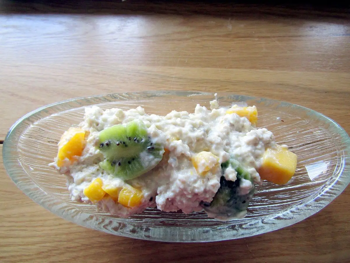 Cuenco de cristal con avena remojada y trozos de mango y kiwi sobre una mesa de madera