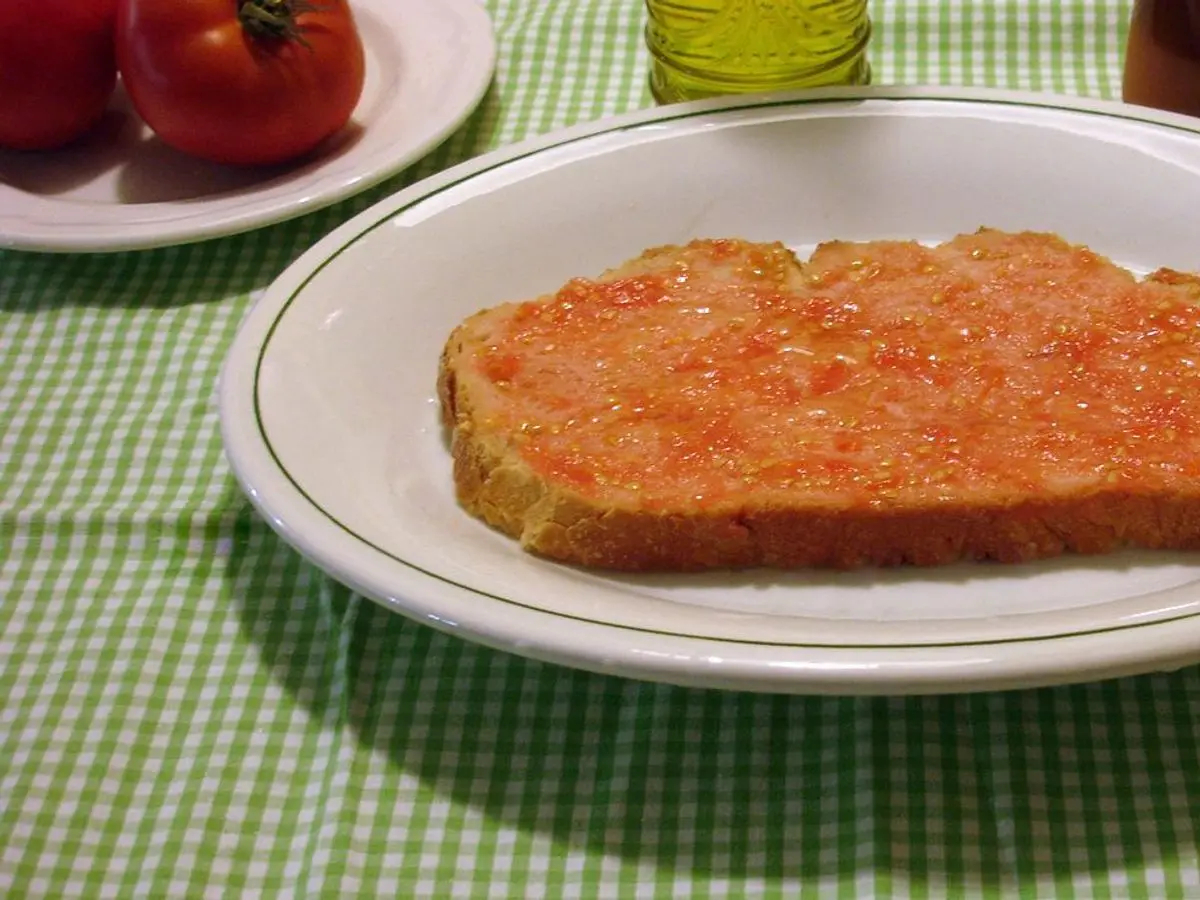 Rebanada de pan rústico untada con tomate y aceite sobre una fuente blanca con mantel de cuadros