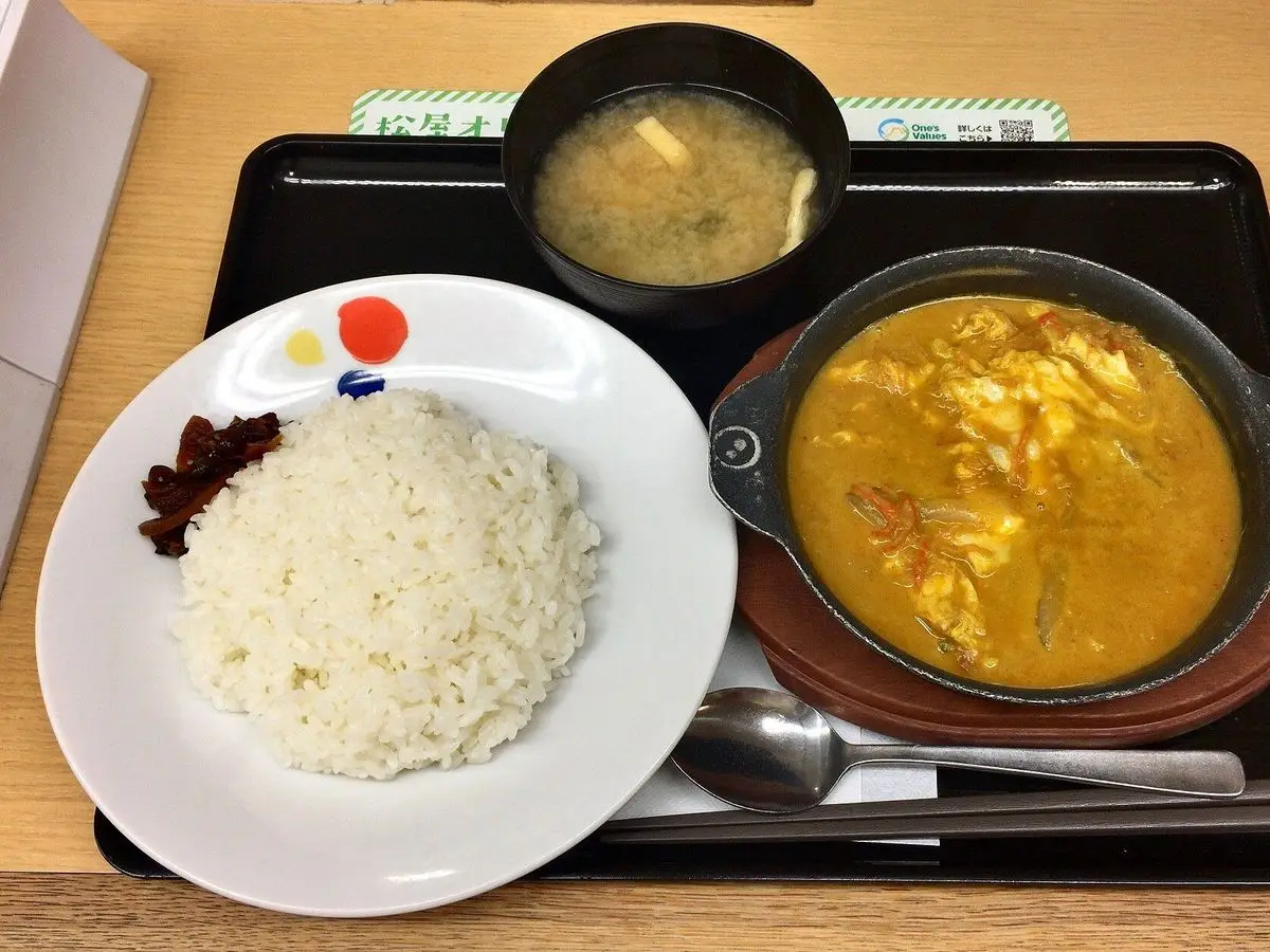 Curry amarillo cremoso con huevo en una cazuela negra junto a un plato de arroz blanco y una sopa