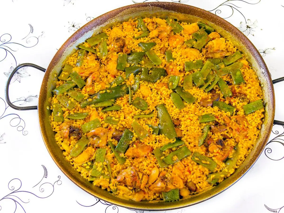 Paellera con arroz dorado, judías verdes planas y trozos de carne vista desde arriba sobre un mantel blanco