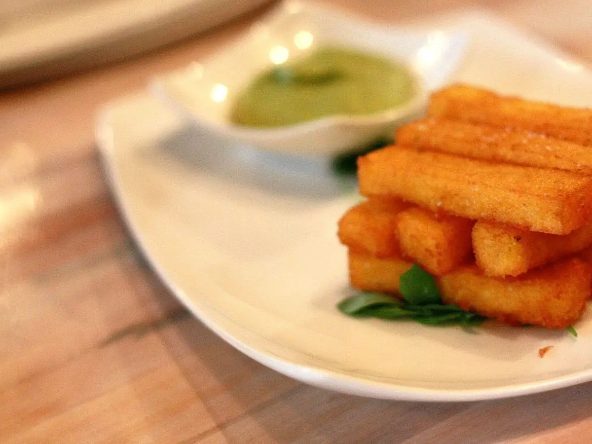 Palitos dorados de polenta frita junto a un cuenco de salsa verde para mojar en un plato blanco