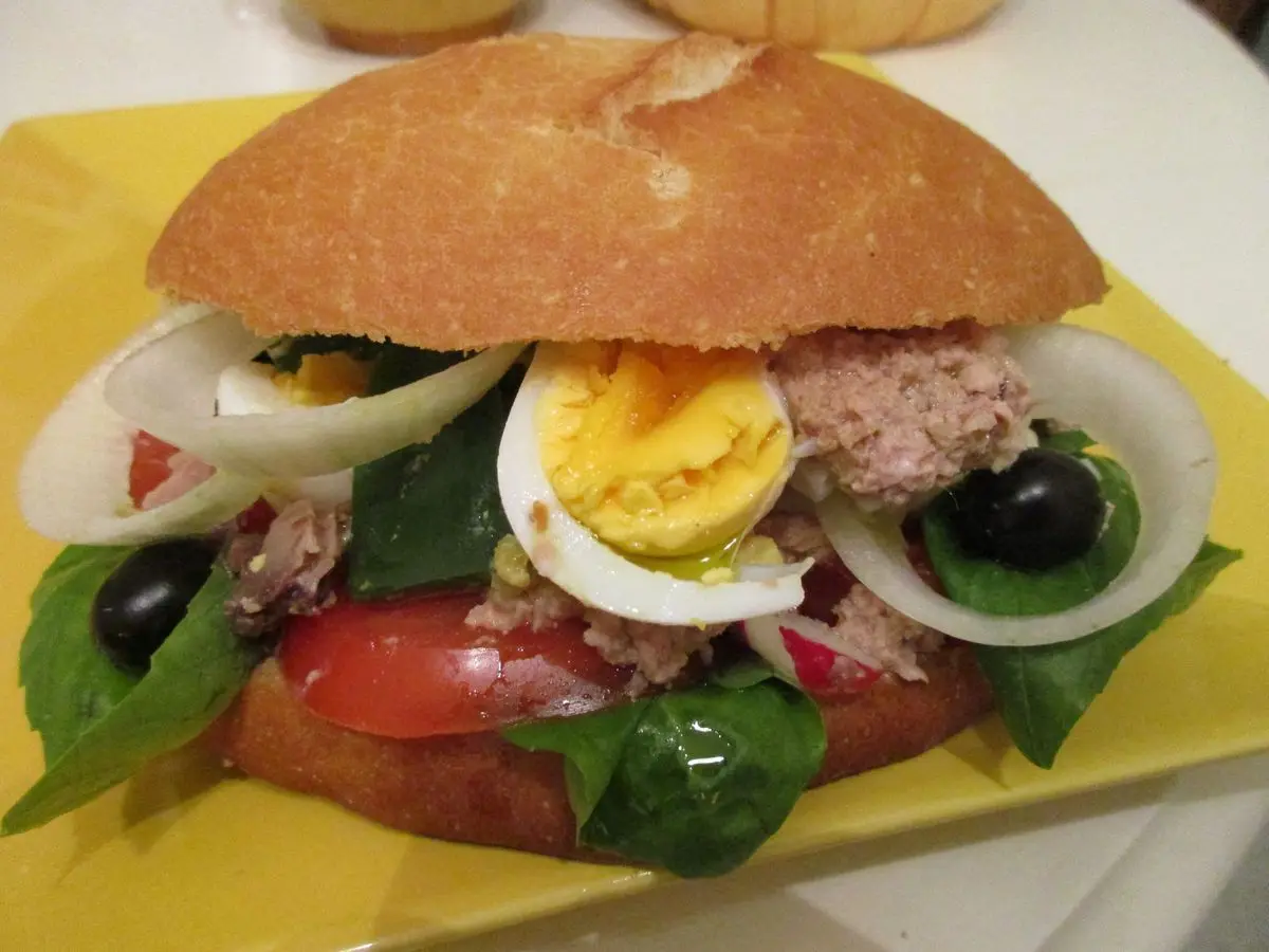 Bocadillo redondo abierto con atún, huevo duro en rodajas, tomate, aceitunas negras y hojas verdes